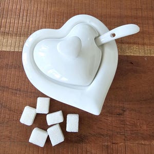 Peut inclure: Un sucrier en céramique blanche en forme de cœur avec un couvercle et une cuillère assortie. Six morceaux de sucre blanc sont éparpillés sur une surface en bois.