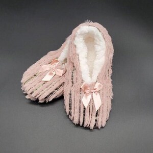 May include: A pair of pink plush slippers with a soft, white interior. Each slipper features a satin bow at the toe and gold-colored stripes. The slippers are designed for indoor wear and provide warmth and comfort.