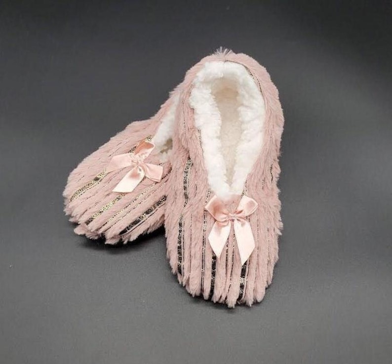 May include: A pair of pink plush slippers with a soft, white interior. Each slipper features a satin bow at the toe and gold-colored stripes. The slippers are designed for indoor wear and provide warmth and comfort.