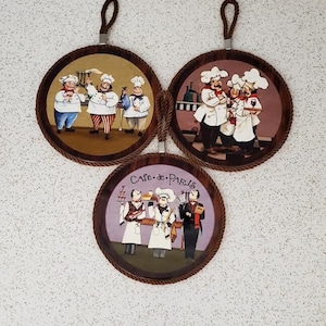 Könnte beinhalten: Drei runde, braune, holzgerahmte Untersetzer mit Seilgriffen. Jeder Untersetzer zeigt eine andere Illustration von drei Köchen in einer Küchenumgebung. Der Text "Cafe de Paris" befindet sich auf einem der Untersetzer.