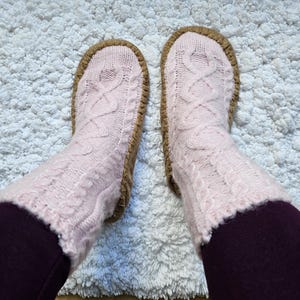 May include: A pair of pink knitted slipper socks with a cable knit pattern and brown suede soles. The socks are worn on a white fluffy rug.