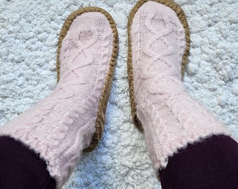 May include: A pair of pink knitted slipper socks with a cable knit pattern and brown suede soles. The socks are worn on a white fluffy rug.