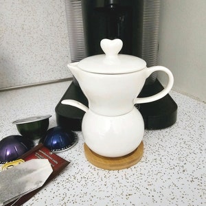 May include: White ceramic coffee pot with a heart-shaped handle and lid. The pot is sitting on a wooden base. There are coffee pods in the background.