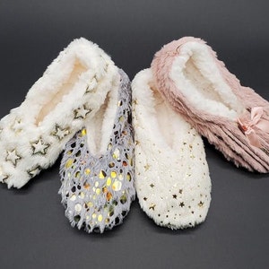 May include: Four pairs of cozy, plush slippers in cream, gray, and pink. The cream slippers have gold star accents, the gray ones have gold dot accents, and the pink ones have a bow. The slippers have a soft, white interior.