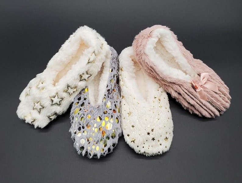 May include: Four pairs of cozy, plush slippers in cream, gray, and pink. The cream slippers have gold star accents, the gray ones have gold dot accents, and the pink ones have a bow. The slippers have a soft, white interior.