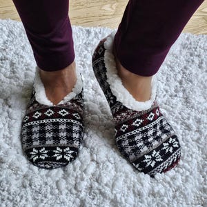May include: A pair of slipper socks featuring a black, white, and maroon fair isle pattern with a geometric design and snowflake motifs. The slippers have a white faux-shearling lining along the collar and are worn with maroon leggings.