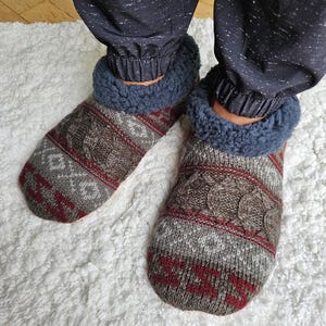 May include: Pair of gray and red patterned slipper socks with a blue and white fleece lining. The socks are worn on a white fluffy rug.