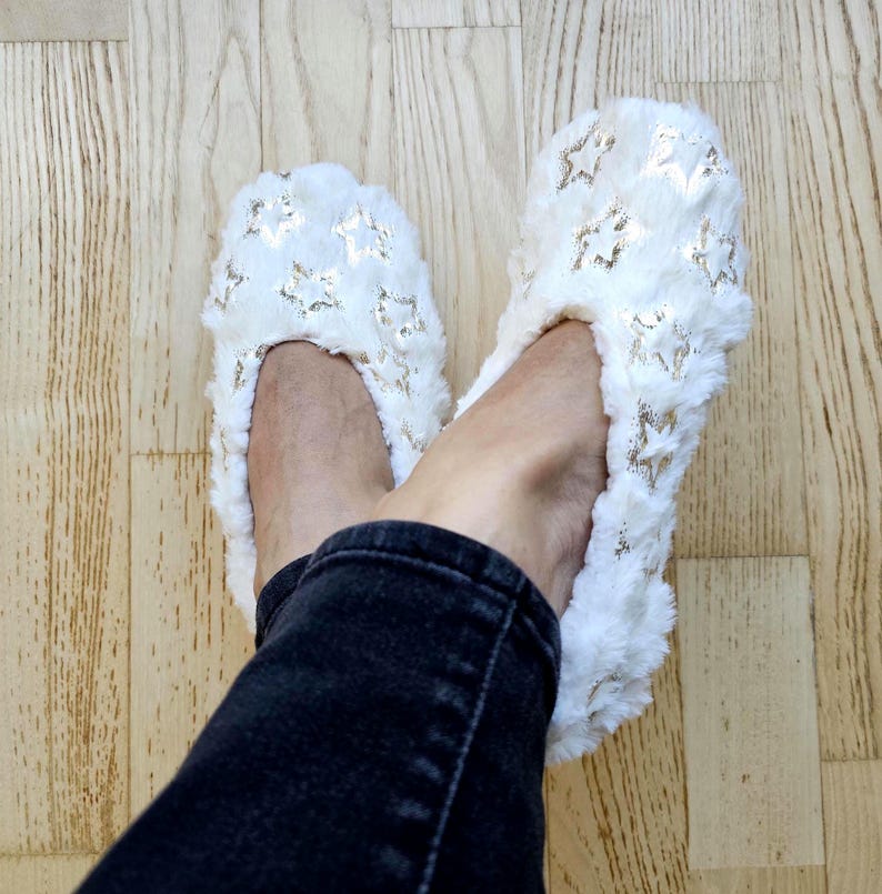 May include: A pair of white, fluffy slippers with gold star patterns. The slippers are on a light wood floor, and the person wearing them is wearing black jeans.