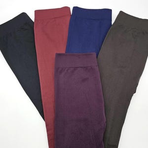 May include: A collection of five pairs of leggings featuring a wide elastic waistband. The leggings are presented in solid colors including black, burgundy, navy blue, dark purple, and charcoal gray, arranged in a fan-like display on a white surface.