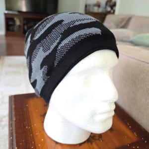 May include: A knit beanie featuring a black and grey camouflage pattern. The hat is constructed from a textured fabric with a solid black band along the bottom edge, designed to fit snugly over the head with a rounded top and a soft, flexible knit.
