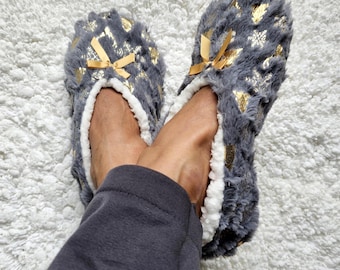 Può includere: Pantofole in peluche grigie e dorate con dettaglio a fiocco. Le pantofole hanno una fodera bianca e soffice e sono decorate con accenti dorati a forma di stella. La persona indossa una tuta grigia e si riposa su un tappeto bianco e soffice.