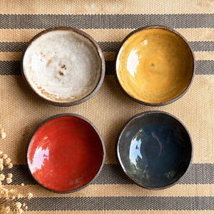 May include: Four small, handmade ceramic bowls in a variety of colors: off-white, yellow, red, and dark blue. Each bowl has a dark brown rim and a glossy finish. The bowls are arranged on a striped beige and brown surface.