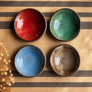 May include: Four small, round ceramic bowls in red, green, blue, and brown. Each bowl has a dark rim and a textured, glazed interior. The bowls are arranged on a striped beige and black fabric.