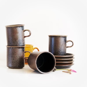 Peut inclure: Ensemble de quatre tasses à café en céramique brune avec un glaçage moucheté. Les tasses sont empilées et l'une est posée sur une soucoupe. Il y a deux soucoupes supplémentaires empilées sur la première soucoupe.