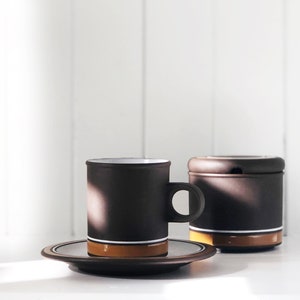 Puede incluir: Una taza de café y un platillo de cerámica marrón con una franja blanca y una franja marrón. La taza tiene un asa y el platillo es redondo. Hay un azucarero a juego de cerámica marrón con tapa.