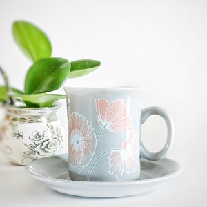 May include: A gray ceramic coffee mug with a floral design in pink and white. The mug has a handle and is sitting on a matching saucer.