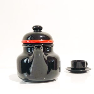 May include: A black teapot with a red band around the lid, and a matching black teacup and saucer set. The teapot has a rounded shape and a spout. The teacup is small and has a handle. The set is on a white surface.