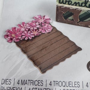 Op de afbeelding: Een gestanste papieren vorm met een bruine rechthoekige basis met horizontale lijnen en een tros roze bloemen aan de bovenrand. Het stuk wordt getoond op een verpakking met de tekst DIES, 4 MATRICES, 4 TROQUELES.