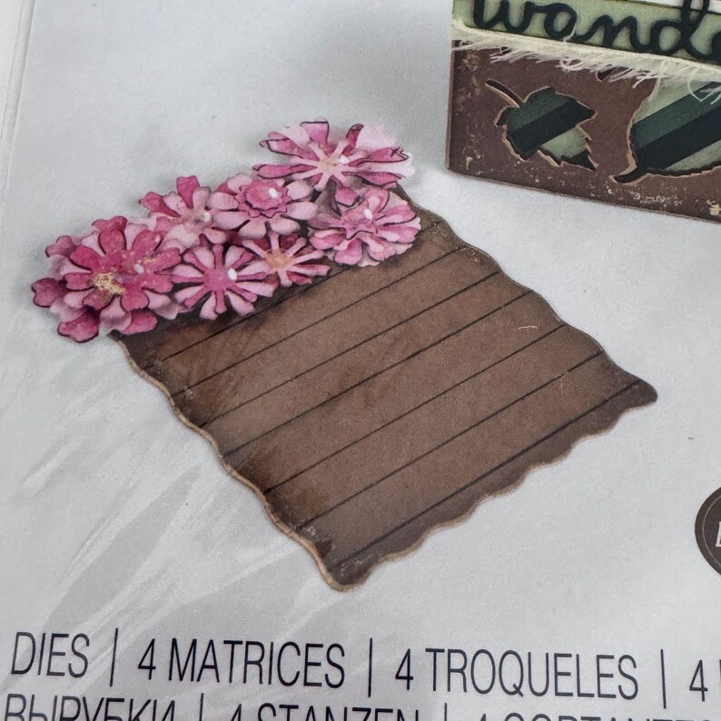 Op de afbeelding: Een gestanste papieren vorm met een bruine rechthoekige basis met horizontale lijnen en een tros roze bloemen aan de bovenrand. Het stuk wordt getoond op een verpakking met de tekst DIES, 4 MATRICES, 4 TROQUELES.