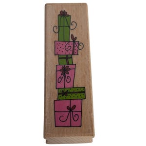 Puede incluir: Un sello de madera con una pila de cajas de regalo coloridas. Los regalos son en tonos verdes y rosas, con lazos y espirales decorativos. El sello es rectangular y está hecho de madera clara.
