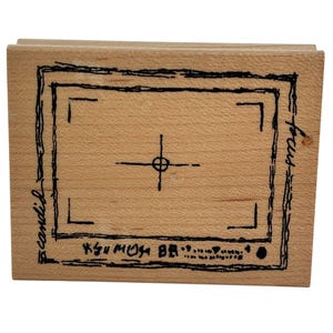 Puede incluir: Un sello de goma rectangular de madera con un diseño en tinta negra de un marco de visor de cámara. El diseño incluye una retícula central, marcas de esquina y las palabras escritas a mano "candid" y "focus" en los bordes laterales.
