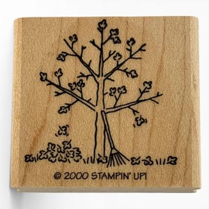 Puede incluir: Sello de madera cuadrado con un diseño de árbol en tinta negra, que incluye hojas, un rastrillo y hierba. El sello tiene el texto "© 2000 STAMPIN' UP!". El sello está hecho de madera clara.