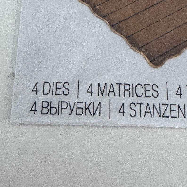 Op de afbeelding: Een wit productverpakkingslabel met de tekst 4 DIES | 4 MATRICES | 4 ВЫРУБКИ | 4 STANZEN gedrukt in een zwart schreefloos lettertype. Een deel van een bruin object met houtnerftextuur en een geschulpte rand is zichtbaar in de rechterbovenhoek.