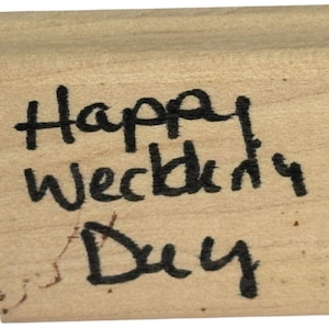 May include: A rectangular wooden stamp with the handwritten text "Happy Wedding Day" in black ink. The stamp has a natural wood grain and a slightly curved top edge.