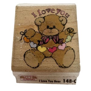 May include: Wooden rubber stamp with a brown teddy bear holding a string of colorful hearts. The text "I love you" is above the bear and "I Love You Bear 148-C" is below the bear.