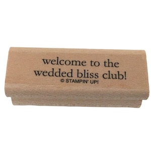 Puede incluir: Un sello de madera rectangular con el texto "welcome to the wedded bliss club!" en negro. El sello también incluye el logotipo "Stampin' Up!". El sello es de color marrón claro y parece sin usar.
