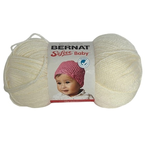 May include: A skein of Bernat Softee Baby yarn in a creamy off-white color. The label features a baby wearing a pink knitted hat. The yarn is soft and suitable for baby projects. The label also includes the word "TESTED".
