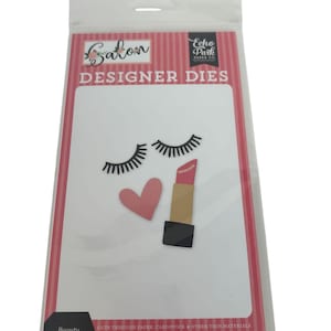 May include: A packet of paper crafting dies labelled "Beauty Die Set". The design includes black eyelashes, a pink heart, and a lipstick. The packet has the text "Salon" and "Designer Dies". Made in America.