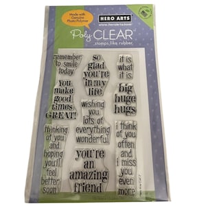 May include: A set of clear photopolymer stamps by Hero Arts on a rectangular backing sheet. The stamps feature various phrases including remember to smile today, so glad you're in my life, it is what it is, big huge hugs, and you're an amazing friend.