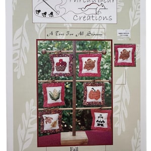 Puede incluir: Una obra de arte enmarcada que presenta un expositor de madera con seis pequeños diseños cuadrados bordados con temática otoñal. Cada diseño está enmarcado en rojo y representa elementos de temporada como una cesta de manzanas, un pavo, maíz, una calabaza y un espantapájaros. La obra se titula "Un árbol para todas las estaciones."