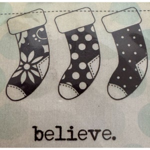 May include: Three black and white Christmas stockings with different patterns hang on a string. The stockings have white fur trim at the top. The word "believe" is printed in black below the stockings.