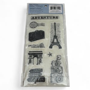 May include: A clear plastic package containing a set of black travel-themed clear stamps. The designs include the Eiffel Tower, a suitcase, a camera, the Arc de Triomphe, a globe, a star, and text reading ADVENTURE and perfect vacation.