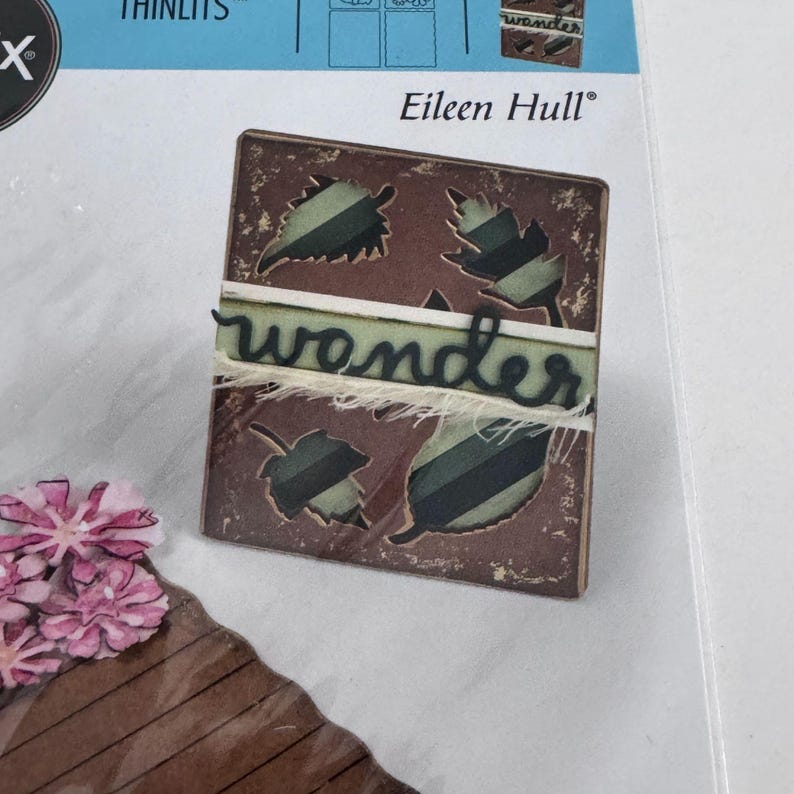 Op de afbeelding: Een vierkant bruin gestanst stuk met vier bladvormige uitsparingen met daaronder groen gestreepte patronen. Een horizontale lichtgroene strook met het woord "wonder" in zwart cursief schrift ligt over het midden, geaccentueerd door gerafelde witte vezels.