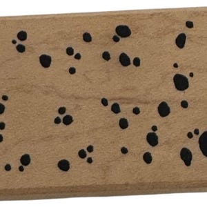 Op de afbeelding: Een rechthoekig houten blok met een lichtbruine afwerking. Het blok is versierd met talrijke zwarte, onregelmatig gevormde vlekken, waardoor een gespikkeld patroon over het oppervlak ontstaat. De vlekken variëren in grootte en dichtheid.