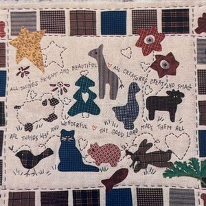 May include: A fabric square with a patchwork design featuring various animal and nature-themed appliqués. The appliqués include a star, sheep, tree, cat, pig, and deer, with embroidered text: "All things bright and beautiful."