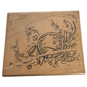May include: A wooden stamp featuring a black line drawing of a French horn with leaves and decorative flourishes. The stamp is square and made of light-colored wood, with the design etched into the surface.