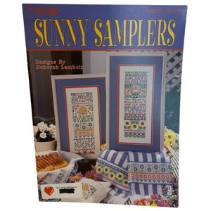 以下が含まれることがあります： デボラ・ランベインによるクロスステッチのデザインが施された「Sunny Samplers」というタイトルの本。表紙には、花柄とアルファベット模様の額入りクロスステッチサンプラーが描かれています。本は青と黄色です。