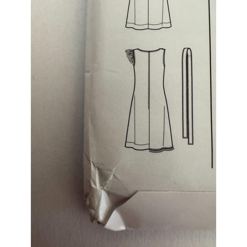 May include: A sewing pattern envelope featuring a black and white line drawing of a sleeveless dress with a ruffled shoulder detail and a separate long fabric tie. The paper envelope is off-white and shows visible creasing and folding at the corner.