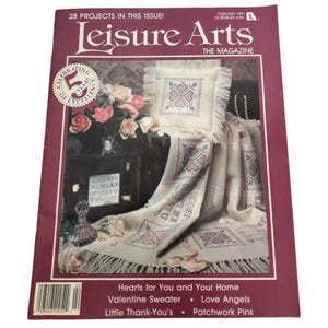 Könnte beinhalten: Titelseite des Leisure Arts Magazins vom Februar 1991. Sie zeigt ein Kreuzstichkissen und eine Decke mit Herzmustern, Rosen und dem Text "Hearts for You and Your Home".