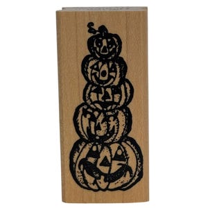 May include: A rectangular wooden rubber stamp featuring a black ink illustration of five stacked jack-o'-lanterns. The pumpkins are arranged vertically, each with a unique carved face, ranging from a small top pumpkin to a large base pumpkin.