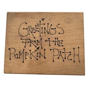 May include: A rectangular wooden sign with the words "Greetings From The Pumpkin Patch" in black handwritten script. The sign has a light brown color and a rustic appearance, perfect for fall decor.