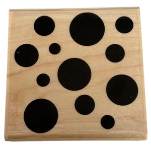 May include: A square wooden rubber stamp featuring a light tan natural wood grain base. The flat surface of the stamp is decorated with a pattern of solid black circles of varying sizes, scattered across the wood in an irregular arrangement.