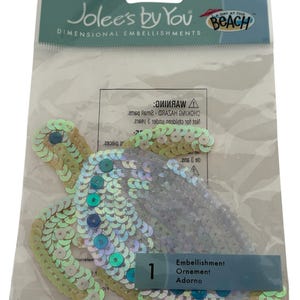 Puede incluir: Un paquete de adornos dimensionales de Jolee's by You. El paquete contiene un adorno de tortuga con lentejuelas verdes, azules y plateadas. El embalaje también tiene el texto "A Day at the Beach".