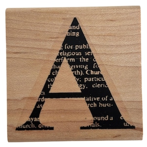 May include: A square wooden block featuring a large, bold, black letter A printed on the surface. The interior of the letter A is filled with a section of black text from a dictionary or book, showing various words and phrases in a serif font.