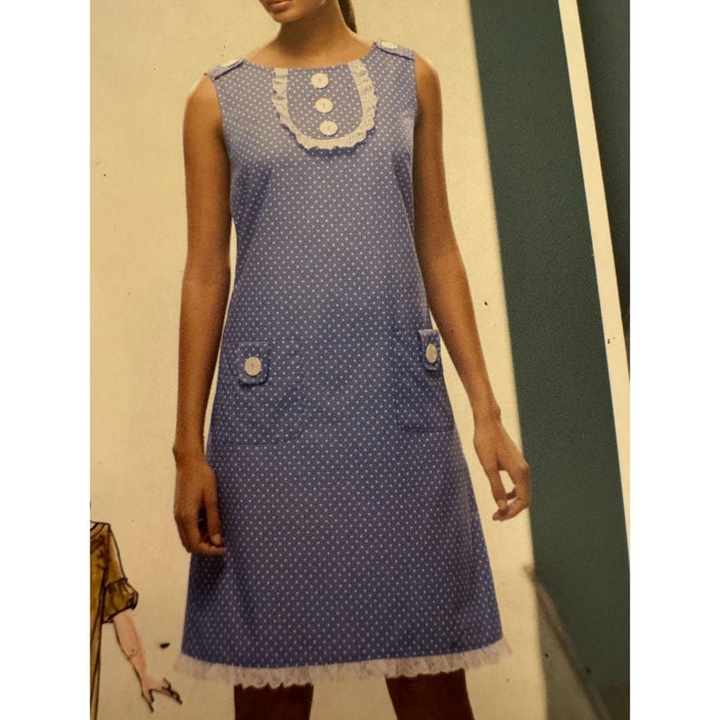 May include: A sleeveless blue dress featuring a white polka dot pattern, a round neckline with a white lace trim, and three white buttons. The dress includes two front pockets, each adorned with a single white button, and a white lace hem.