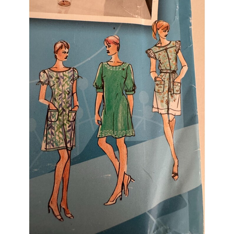 May include: A sewing pattern envelope featuring three illustrations of knee-length dresses. The left dress has a leaf print and patch pockets. The center dress is solid green with short sleeves. The right dress has a floral print and a waist tie.